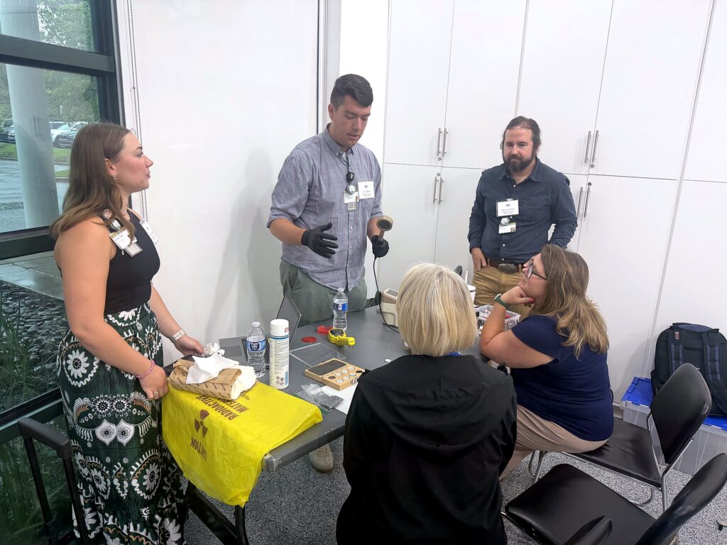 A facilitator wearing a glove demonstrates a handheld radiation detector beside a yellow radioactive-materials bag as teachers observe.