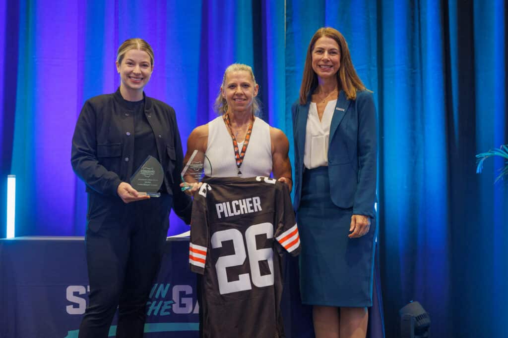 Three educators hold a glass trophy and a Cleveland Browns jersey at a Stay in the Game! award ceremony.