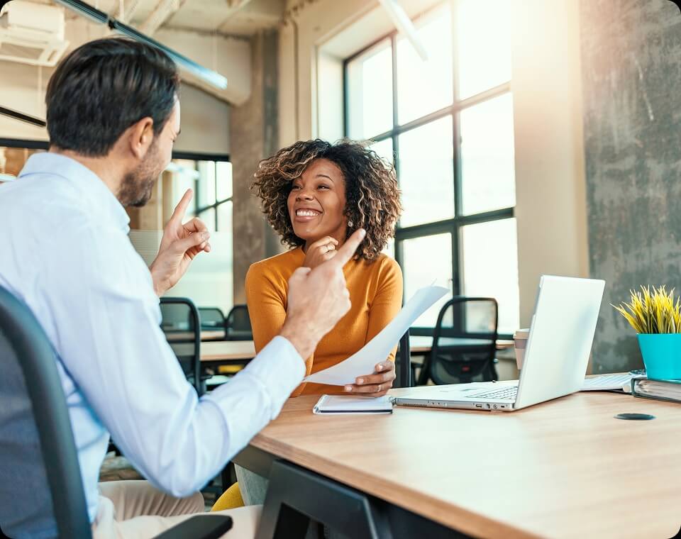 Two colleagues sitting at a table smiling and talking while holding a sheet of paper