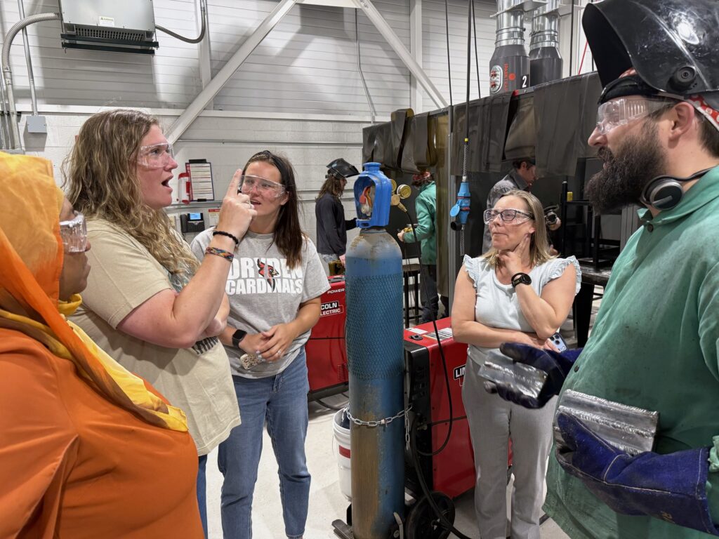 A teacher in safety glasses talks with a welding instructor beside a welding bay during a Michigan industry externship.
