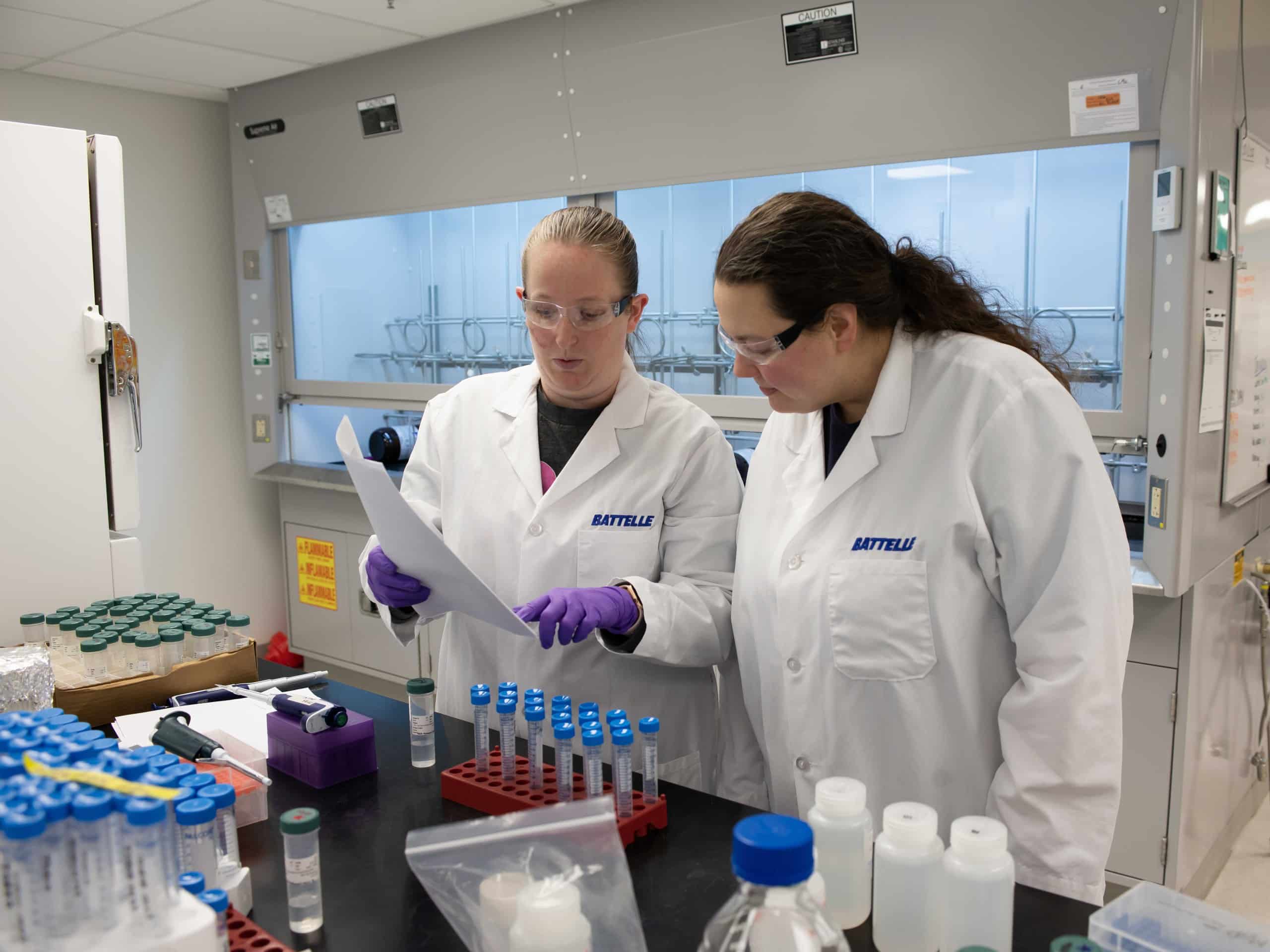 Two Battelle scientists in lab coats and safety glasses review a document together at a laboratory bench lined with sample tubes.