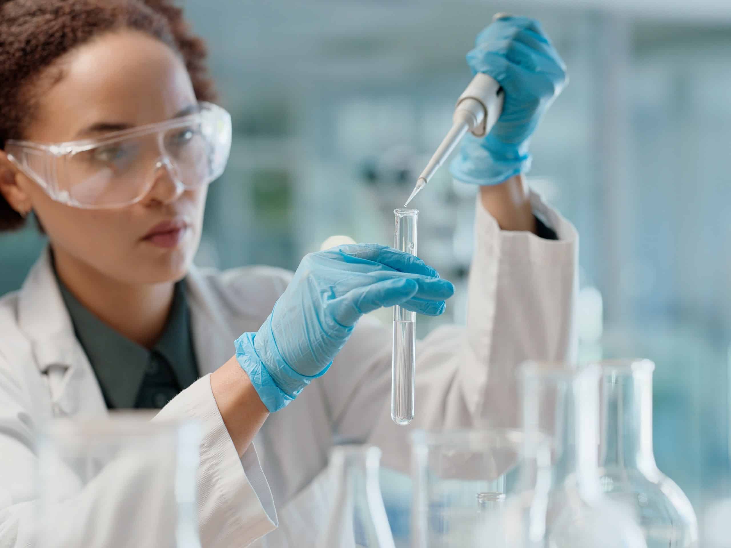 A scientist in a lab coat, safety goggles and blue gloves uses a pipette to transfer liquid into a test tube.