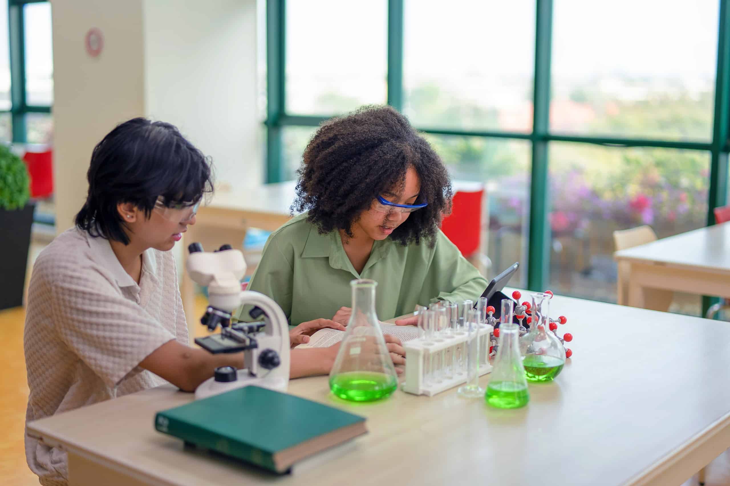 Two high school students in safety glasses study at a science lab table with a microscope, a notebook and flasks of green liquid.