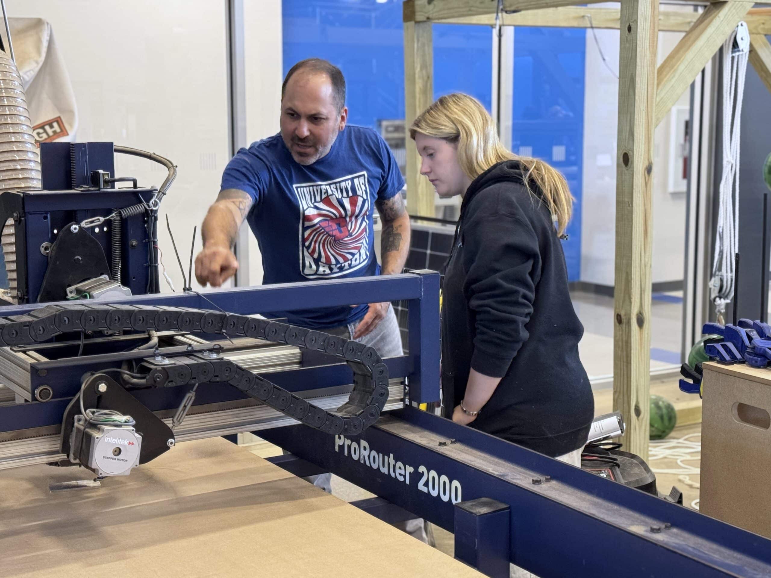 An instructor points out a detail on a ProRouter 2000 CNC machine to a student in a fabrication lab.