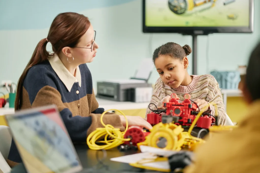 A teacher seated at a table while a young girl works on a robotic device