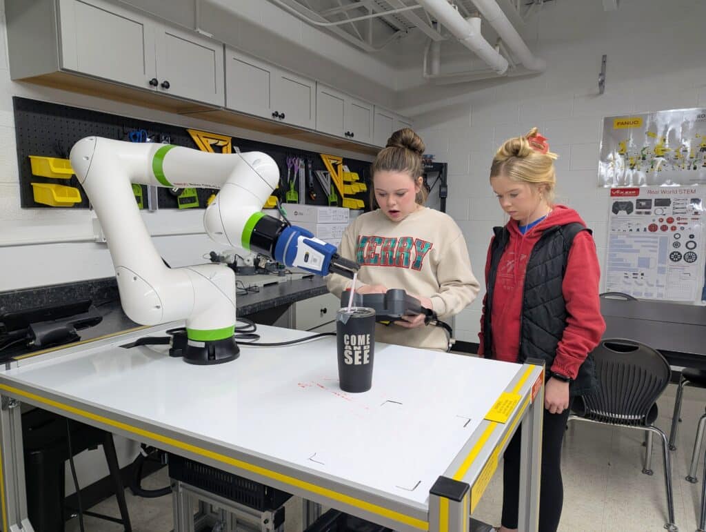 Two students operate a FANUC robotic arm using a handheld controller in a robotics classroom.
