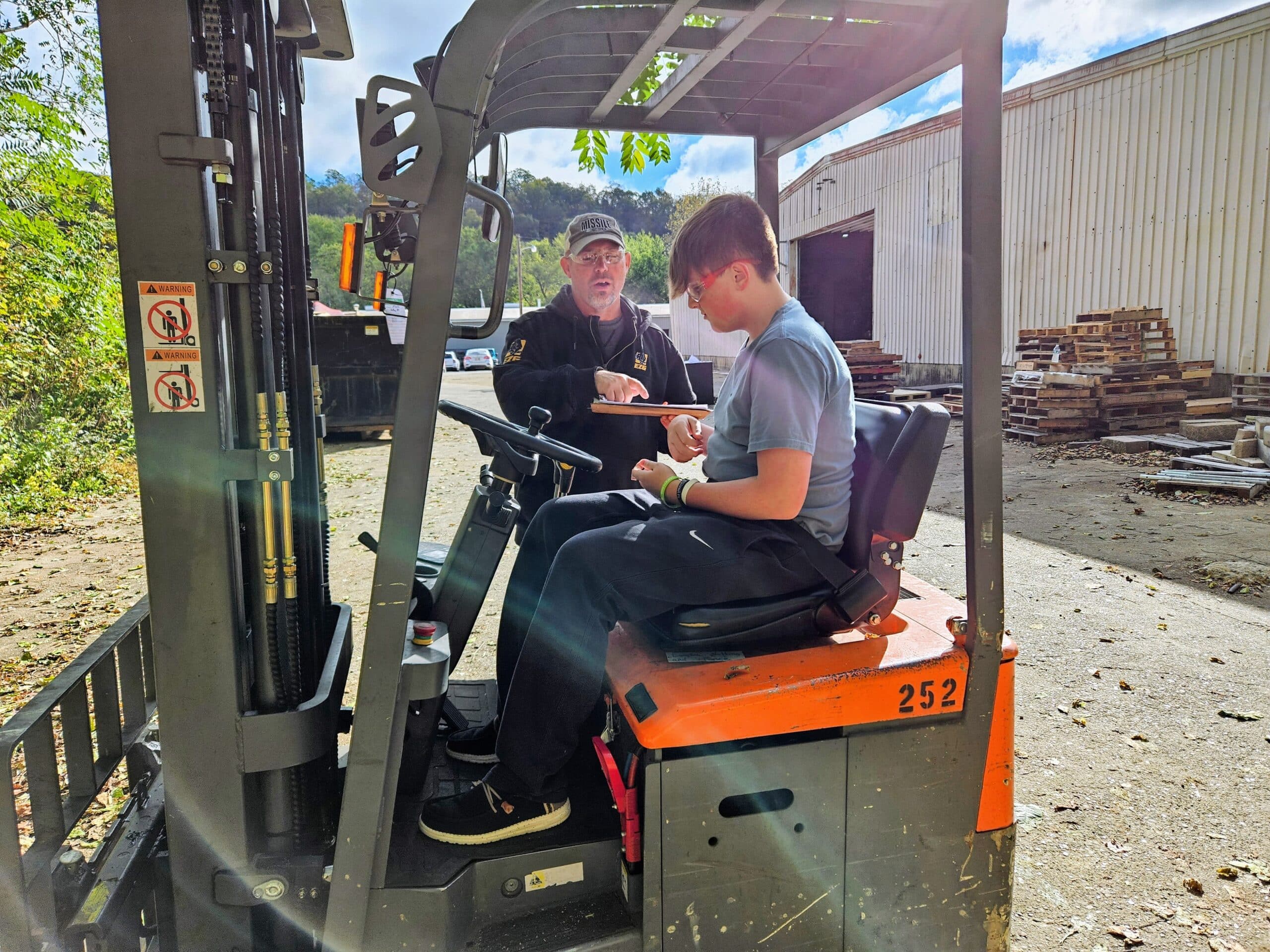An instructor guides a student seated on a forklift during a hands-on industry training session at an outdoor equipment yard.