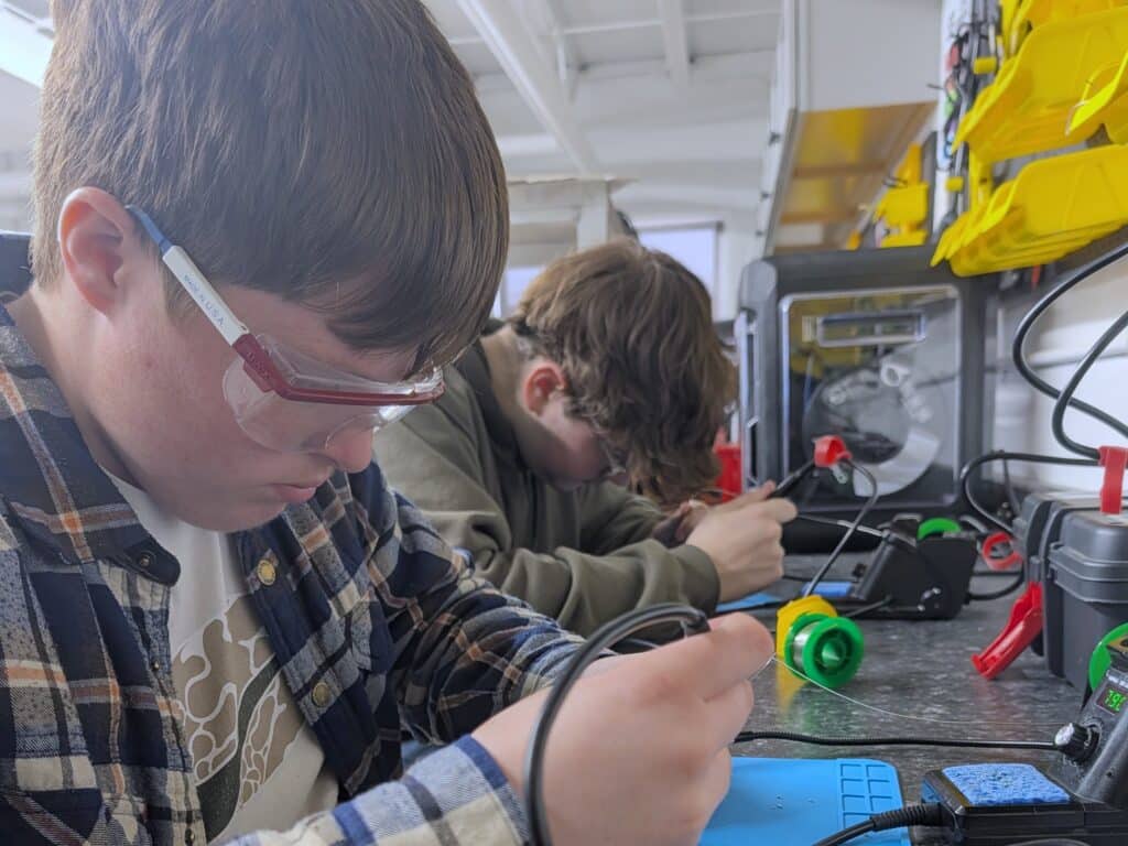 A student wearing safety glasses solders an electronic component at a workbench while another student works nearby in a fabrication lab.